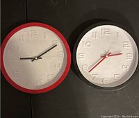 Two round wall clocks laid side-by-side on a flat surface. Left clock has a red rim and a crack; right clock has a white rim. Both clocks have raised numbers and three hands.