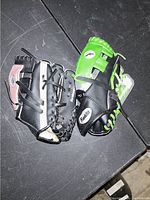 Two kids baseball gloves, one black with pink accents, one black and green with white and green laces, placed on a dark surface.