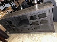 Front angle view showing wooden grey TV stand with two glass paneled doors and central shelf, resting on concrete floor.