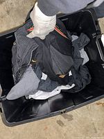 Photo showing various mixed men's clothing items in dark colors in a black plastic tub, mainly tops and jackets.