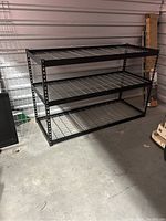 Three-tier black metal shelving unit with wire mesh shelves and adjustable metal frame photographed indoors on a concrete floor.