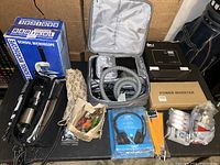 Overview photo of lot items arranged on table: boxed microscope, travel bag with metallic containers, headphones, power inverter, pencils, light bulbs, small bags, patterned pouch.