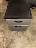 Front and top view of small two-drawer file cabinet showing beige metal drawers with black oval handles and black textured top surface.