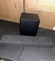 Front and top view of black Vizio soundbar with visible control icons, placed in front of a black square subwoofer on a dark surface with cardboard background.