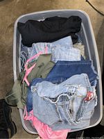 Top-down photo of a plastic bin filled with folded women's clothing, showcasing denim jeans and various colored tops including black, olive green, and pink garments.