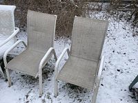 Two white alloy frame lawn chairs with beige mesh fabric seats and backs placed outside on snow-covered ground, showing outdoors use and need for cleaning.