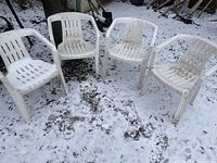 Four white plastic stackable arm chairs outdoors on snow-covered ground.