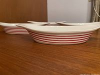 Side view of one white oval casserole dish with horizontal red stripes and integrated handle on a wooden surface.
