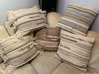 Five hand woven throw pillows arranged on a beige chair, showing neutral colors and textured woven stripes with tassels and tufted patterns.