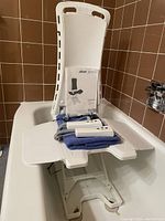 Full view of the white bath lift installed in a tub showing seat, backrest, and hand controls on a towel.