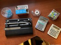 Lot contents on wooden table including calculators, 3-hole punch, tape dispenser, box of staples, containers with tacks and push pins.