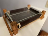 Side perspective showing the wooden serving frame with corner posts and horizontal handles supporting the brown ceramic casserole dish with glass interior.