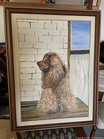 Full view of framed oil painting showing the brown curly-haired dog sitting indoors by a window and brick wall.