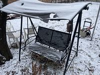 Front side view of the lawn swing showing the torn fabric canopy with snow accumulation, metal frame, and mesh seat and backrest under light snow cover.