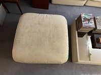 Top view of the beige fabric square ottoman with rounded corners on carpeted floor.