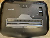Close-up image of the top panel showing brand Staples, paper feed slot, control switch, and instruction icons.