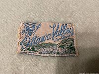 Close-up photo of the embroidered fabric label showing 'Ottawa Valley Woolens' in blue and green stitching with a scenic design.