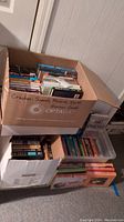 Overview of cardboard boxes and plastic bin filled with paperback books including romance and murder mystery genres