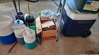 Wide shot showing large wheeled cooler, insulated jugs, plungers, and cardboard boxes of camping cookware all stored on garage floor.
