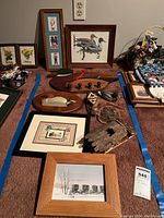 Wide view showing multiple framed art pieces, two wooden duck decoy plaques, tree bark mounted art, and a wooden duck hanger with hooks on carpet.