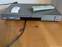 Front view of Samsung DVD Player with remote control on top, showing model and function buttons.