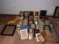 Overview of assorted empty wood and metal photo frames arranged on carpet floor, sizes up to 8x10, varying colors and finishes.