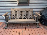 Front view of the wooden outdoor glider chair with decorative backrest and slatted seat, showing full width and armrests.