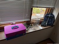 Purple Caboodles storage container with handle, navy blue swing backpack, two wallets, and various jewelry pieces on a windowsill.