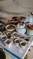 Various ceramic planters arranged on a table in a basement setting with ramp to ground level. Includes terracotta pots, glazed pots, a white planter with removable top, and a wooden planter box containing small ceramic cups.