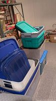 Photo showing stacked plastic storage bins in blue, white, teal with lids on top and sides, set on carpet floor in basement.