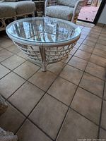 Photo of round rattan coffee table with a clear glass top in a tiled room with other rattan furniture visible.