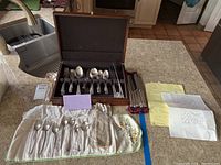 View of the overall lot showing the wooden case open with silverplate flatware organized inside, hand-embroidered sleeve with extra spoons, and accompanying notes.
