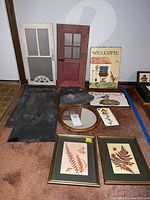 Photo shows two framed pressed flower artworks, three handpainted slates with welcome and garden themes, two blank large slates, two small decorative wooden doors, and one round mirror with a wooden frame all arranged on carpet.