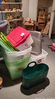 Stack of various plastic storage containers and laundry baskets, including clear, green, and pink lids, displayed on carpeted floor.