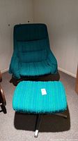 Wide angle photo showing the vintage teal fabric swivel chair and matching footstool placed on a carpeted floor against a wall. Shows overall condition and style.