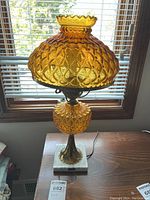 Full view of the lamp showing amber textured glass shade and body, on a square white marble base, placed on a wood surface near a window.