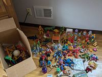 Group of various vintage toys laid out on hardwood floor including action figures, small figurines and a cardboard box with additional items