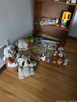 Group of vintage plush toys sitting on floor beside wooden shelf. Playskool flashlight with coloring books and puzzle atop shelf, Oscar Mayer small figurines and plastic toy hot dogs in front.