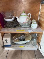 Cabinet shelf containing floral tea set including teapot and sugar bowl, glass bowls and plates, decoupaged glass plates, and a white ceramic baking dish in original packaging.