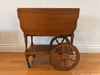 Side view of antique walnut tea wagon with drop leaf extended, decorative large wheel and two small caster wheels, and lower shelf visible.