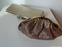 Gold hard case clutch with textured finish and rhinestone-clasp, alongside bronze beaded ruched fabric clutch with gold-tone metal frame and clasp, displayed on white surface.