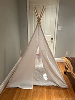 Photo of a children's collapsible teepee playhouse from front, showing fabric walls and wooden support poles tied at the top.