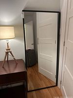 Full length mirror standing upright against a white door, black framed with visible edge damage at the top left corner of the frame.