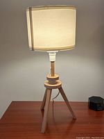Lamp turned on, showing beige drum shade with soft light and natural wood tripod base on wood surface.