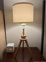 Lamp illuminated showing beige shade and wooden tripod base on a wooden surface with a clock nearby.