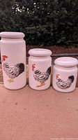 Three white ceramic canisters of varying sizes with embossed chicken designs, displayed side by side outdoors.