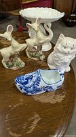 Five vintage ceramic figurines displayed: two deer, one cat, a Dutch clog planter, and a white pedestal dish with swan bases on a wood table.