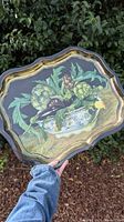 Full view of the decorative tin tray with scalloped gold edges and vegetable design held outdoors.