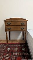 Front view of a medium brown wooden nightstand with two drawers and metal handles, standing on four legs over a patterned rug.
