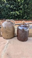 Two stoneware jugs displayed outdoors on a cloth with greenery in background. Beige large speckled jug with handle and smaller dark brown glazed jug with cork stopper.